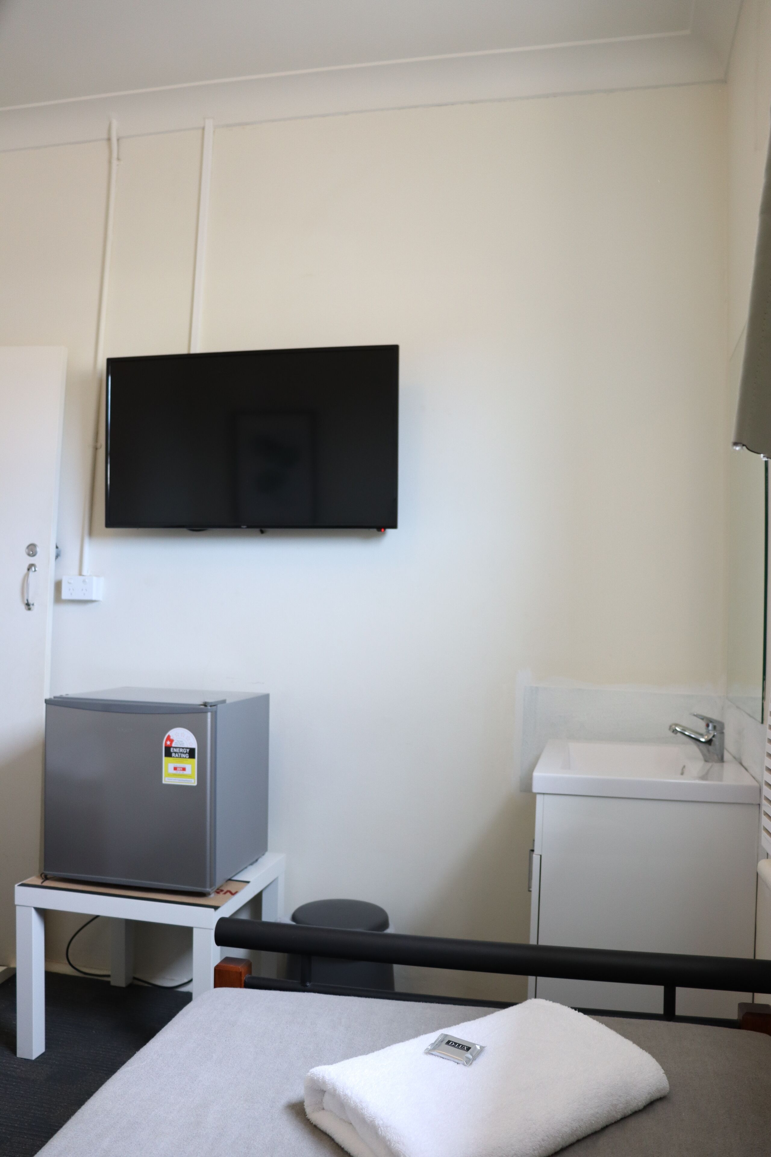 Habitación de hotel con cama, una mesita con refrigerador, televisor montado en la pared, lavamanos y espejo.