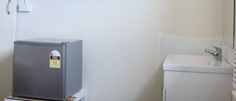 Habitación de hotel con cama, una mesita con refrigerador, televisor montado en la pared, lavamanos y espejo.