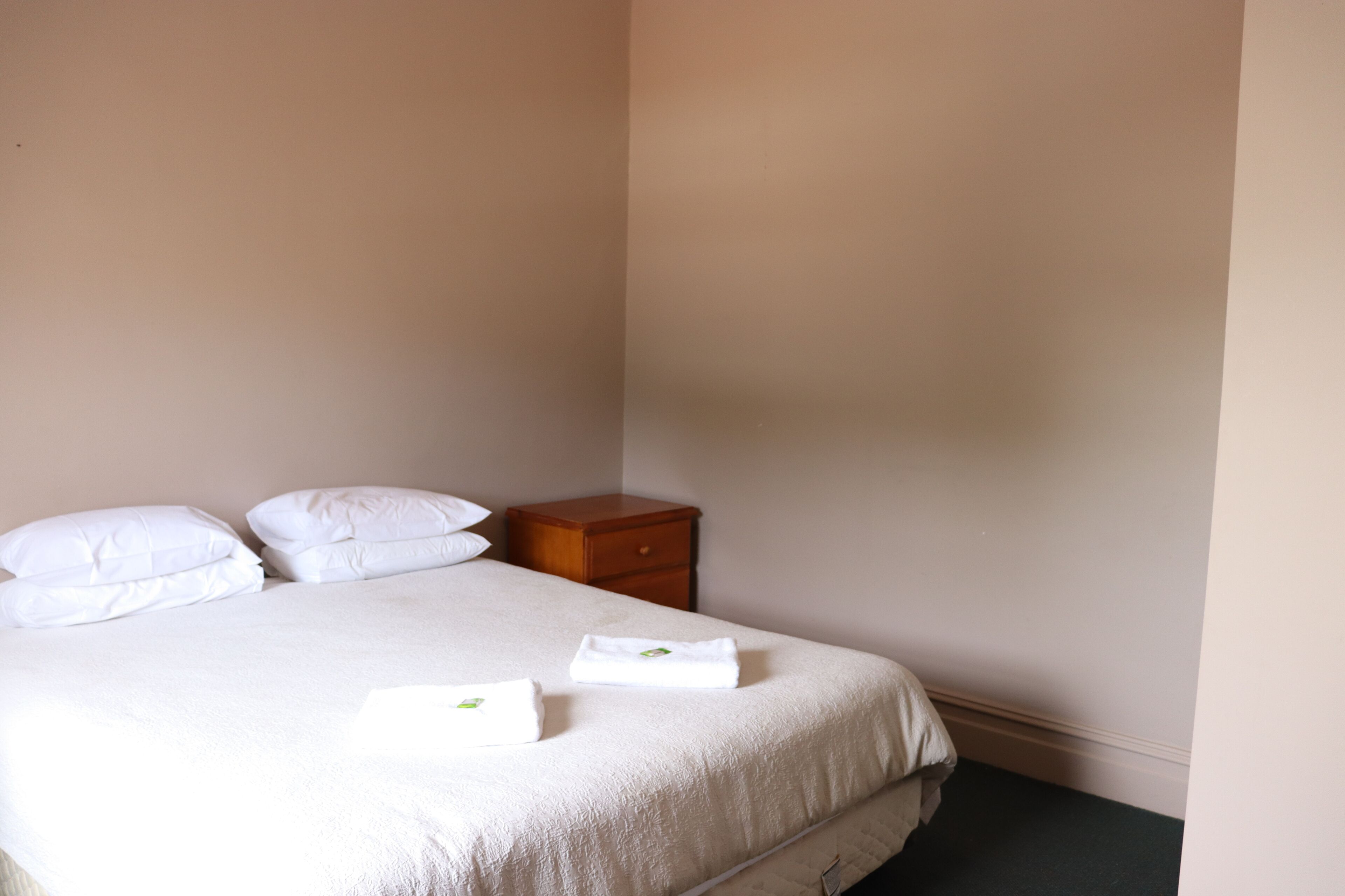 A neatly made bed with white linens and pillows, a wooden nightstand with a drawer, and a plain wall.
