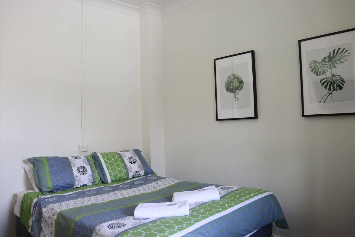 A neatly made bed with a green and white patterned bedspread, two pillows with geometric designs, and two towels folded on the bed.