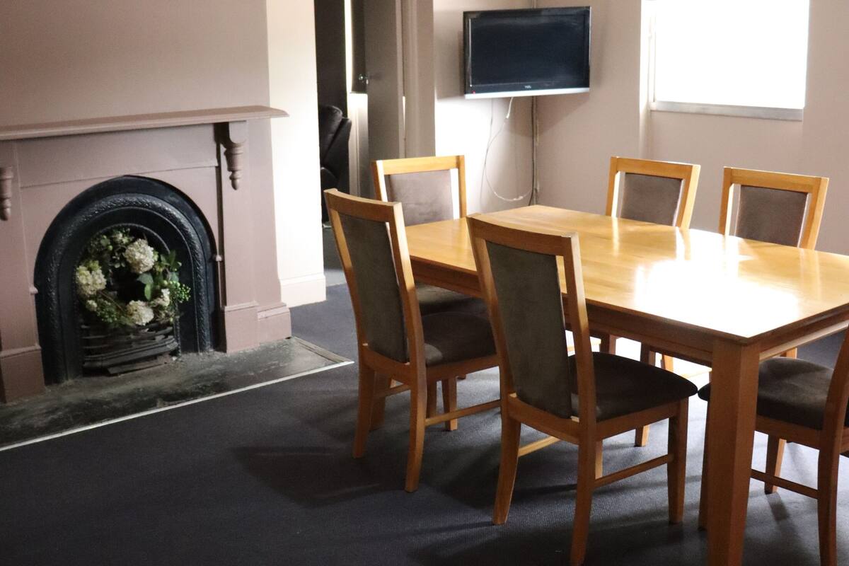 A dining area with a rectangular table and six chairs, a fireplace with a flower arrangement, a television on the wall, and a window providing natural light.
