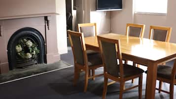 Un área de comedor con una mesa rectangular y seis sillas, una chimenea con un arreglo floral, un televisor en la pared y un ventanal que proporciona luz natural.