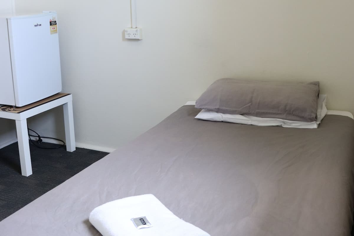 A hotel room with a bed, a white bedside table, a white refrigerator, and a framed plant on the wall.