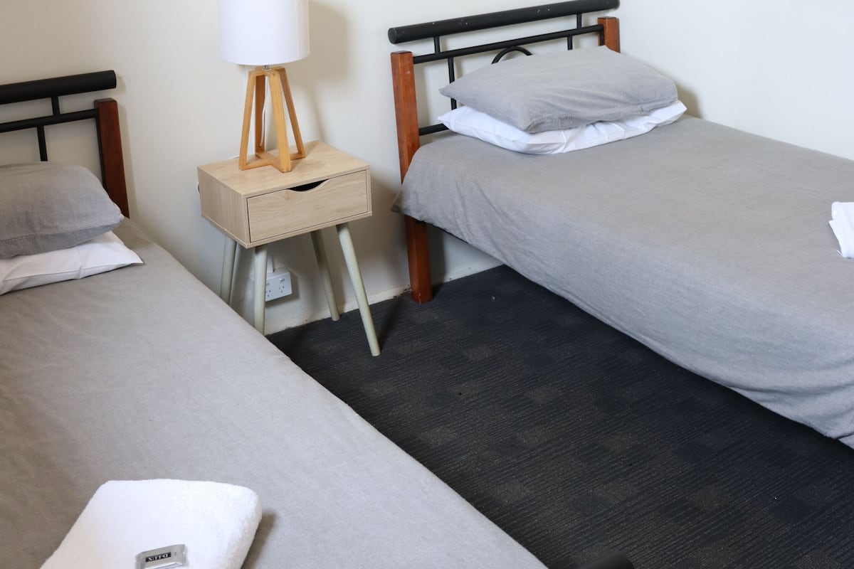 A hotel room with two single beds, a bedside table with a lamp, a framed plant on the wall, and a neatly folded towel on the bed.