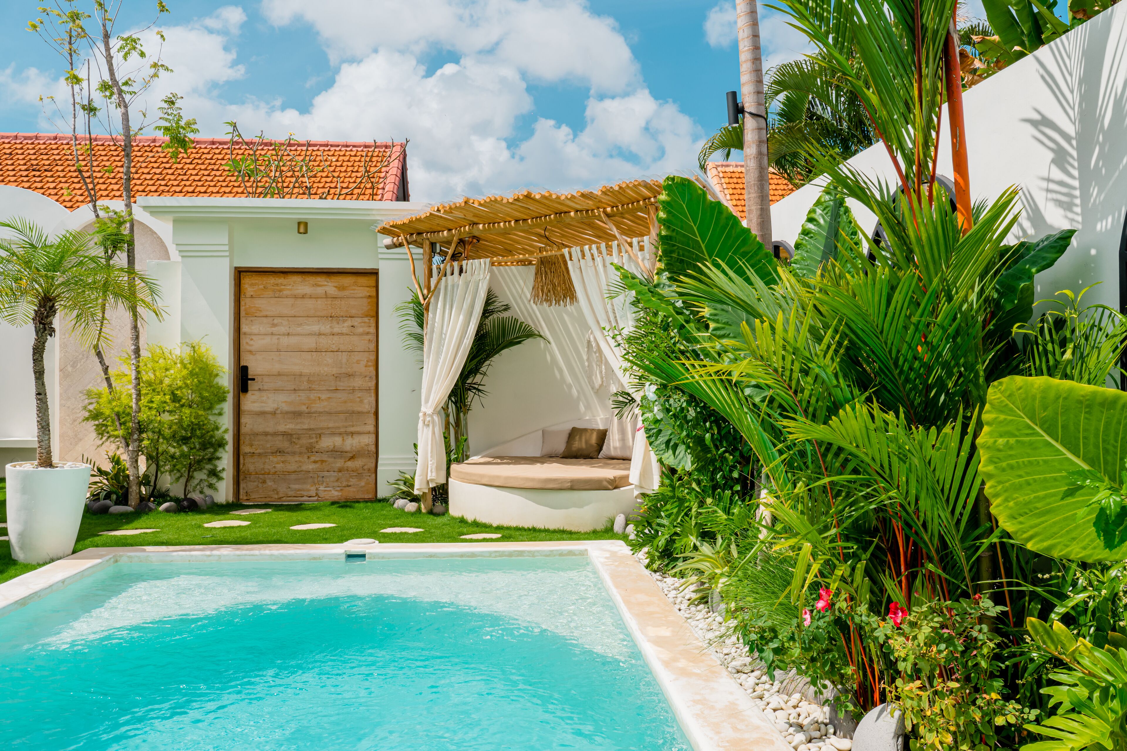 Piscina rodeada por vegetação exuberante, porta de madeira e área de estar coberta com toldo branco.