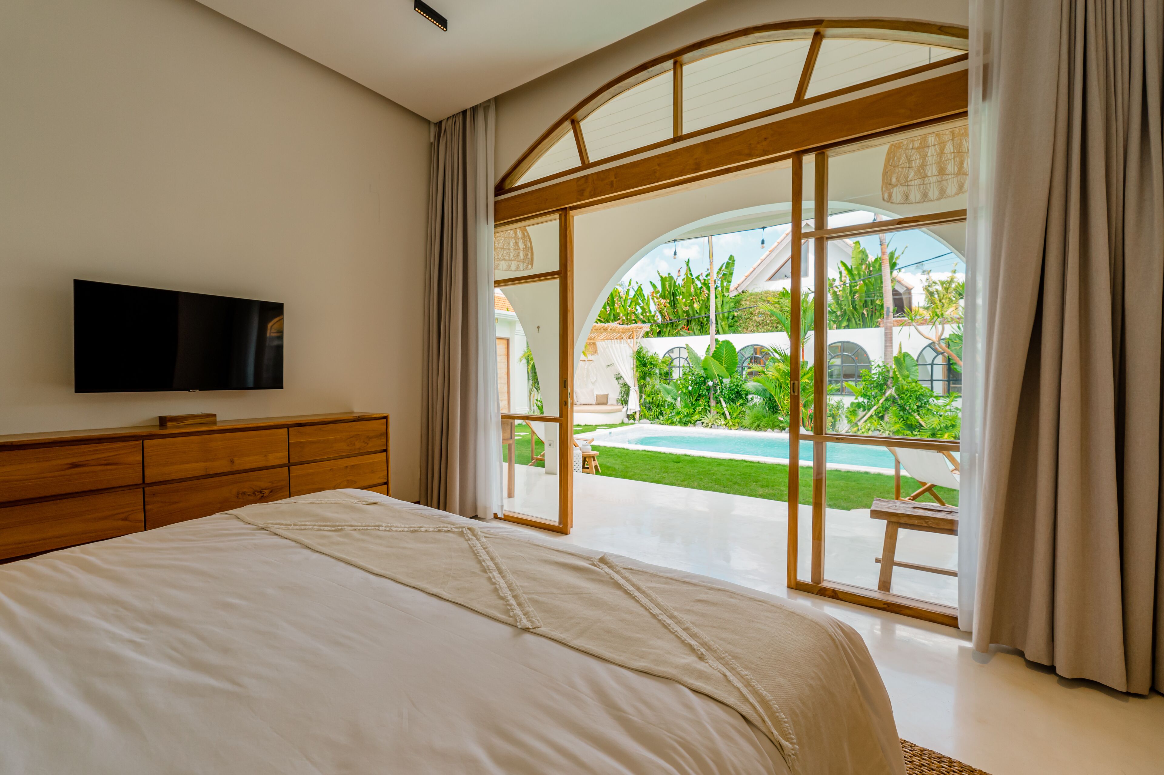 A bedroom with a large bed, a TV mounted on the wall, and a view of a swimming pool and garden through a glass door.