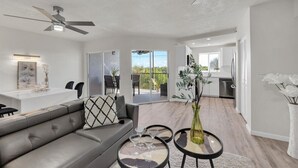 A modern living room with a grey sofa, a dining table, and a ceiling fan. - Jupiter Bay Resort Condo, steps to the beach! Comfortably sleeps family of 4! (Jupiter)