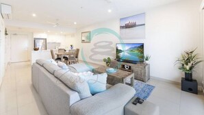 A modern living room with a grey sofa, a wooden coffee table, a flat-screen TV, and a ceiling fan. - Zen Sun City - 2 BR Cozy Apt Ocean View (Darwin)