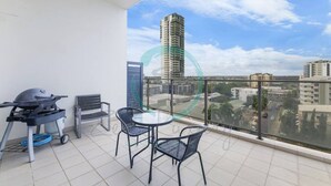 A balcony with a table and chairs, a grill, and a view of a tall building and surrounding area. - Zen Sun City - 2 BR Cozy Apt Ocean View (Darwin)