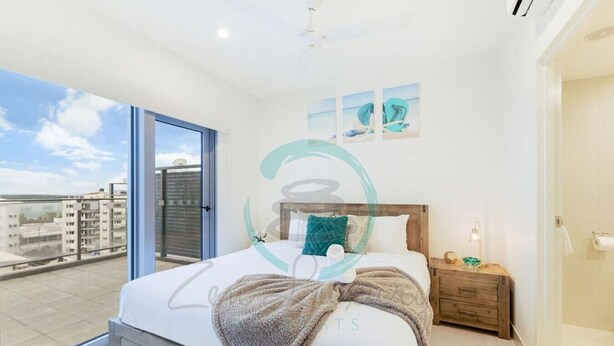 A modern bedroom with a large bed, a wooden nightstand, a bedside table with a lamp, and a view of the city through a large window. - Zen Sun City - 2 BR Cozy Apt Ocean View (Darwin)