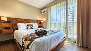 Une chambre d’hôtel avec un grand lit, une tête de lit en bois, deux tables de chevet avec des lampes, une fenêtre avec des rideaux légers et un appareil de climatisation.