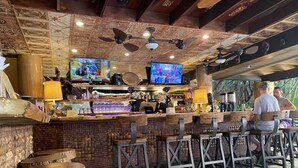 Un coin bar rustique avec des tabourets de bar en bois, des ventilateurs de plafond et un écran de télévision diffusant une émission.