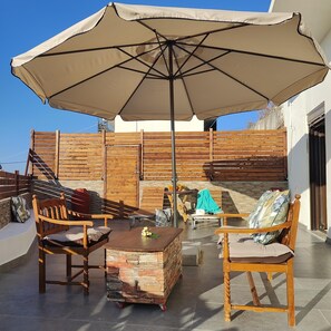 Un espace patio aménagé avec une table en bois, des chaises, un banc en pierre et un grand parasol.