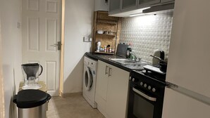 Pieni ja käytännöllinen keittiö, jossa on pyykinpesukone, liesi, pesuallas ja pieni hylly, jossa säilytetään keittiötarvikkeita.
