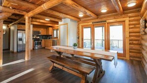 Un interior espacioso de cabaña de madera con una gran mesa de comedor, una zona de cocina y un balcón con puertas de vidrio.