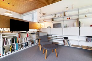 Una biblioteca moderna con una televisión de pantalla plana, estanterías, un sillón gris y techo de madera.