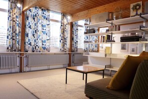 Una habitación con un sofá, una mesa, estanterías llenas de libros y objetos decorativos, y una ventana con cortinas estampadas.