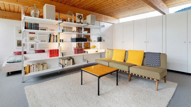 Een moderne woonkamer met een houten plafond, witte kasten, een bank met gele kussens en een salontafel.