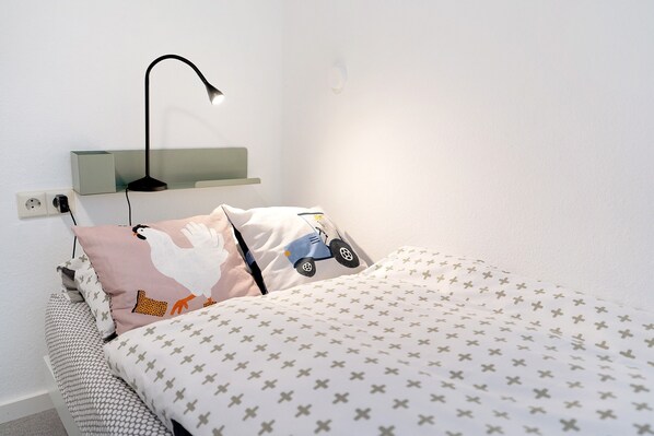 A neatly made bed with patterned bedding, a wall-mounted lamp, and a pillow with a vehicle design.