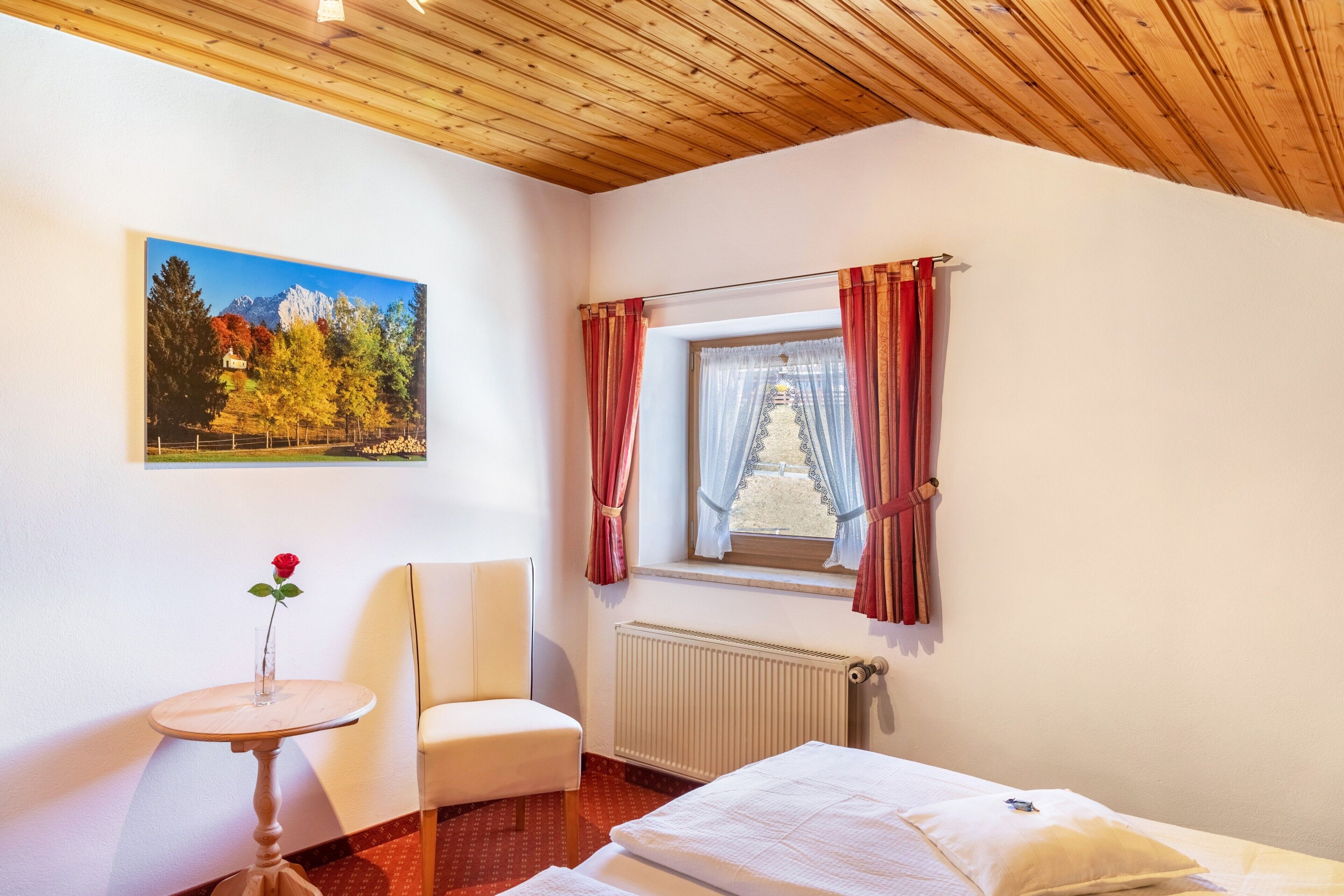 Quarto com cama, uma cadeira, uma mesinha com uma rosa, janela com cortinas e um quadro na parede.