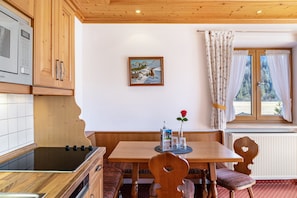Une cuisine avec des meubles de rangement en bois, un four à micro-ondes, une cuisinière, une table à manger avec des chaises, un tableau accroché au mur et une fenêtre avec des rideaux.