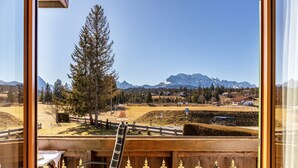 A view from a window with a wooden railing, a table, and a chair, overlooking a mountainous landscape with buildings and a clear sky.