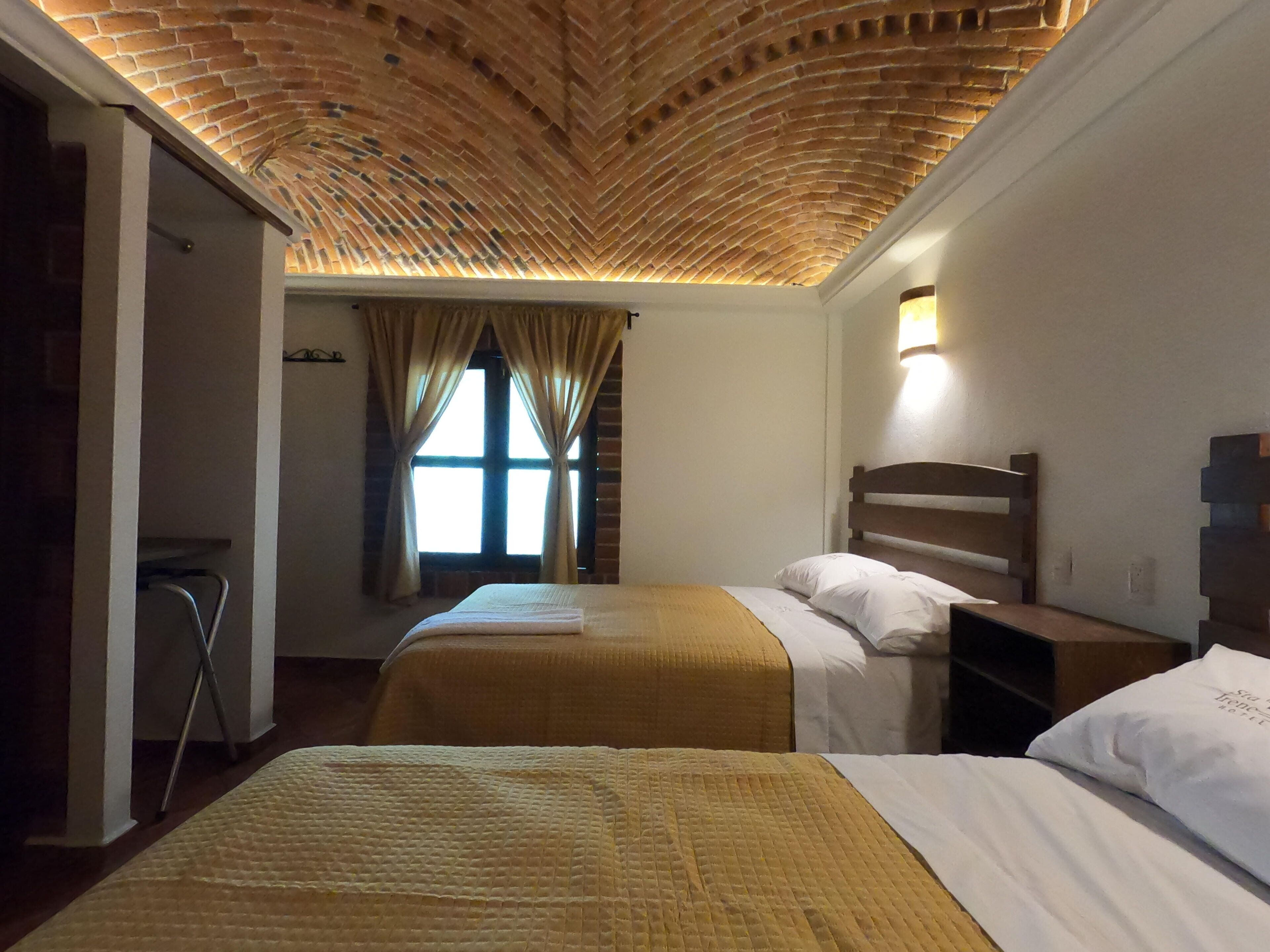 A room with two beds, a wooden ceiling, a window with curtains, and a nightstand with a lamp.
