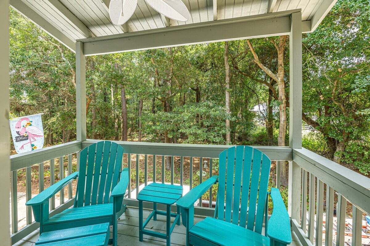 Een overdekt terras met twee turquoise Adirondack-stoelen en een kleine tafel, omgeven door een houten leuning en weelderige begroeiing.