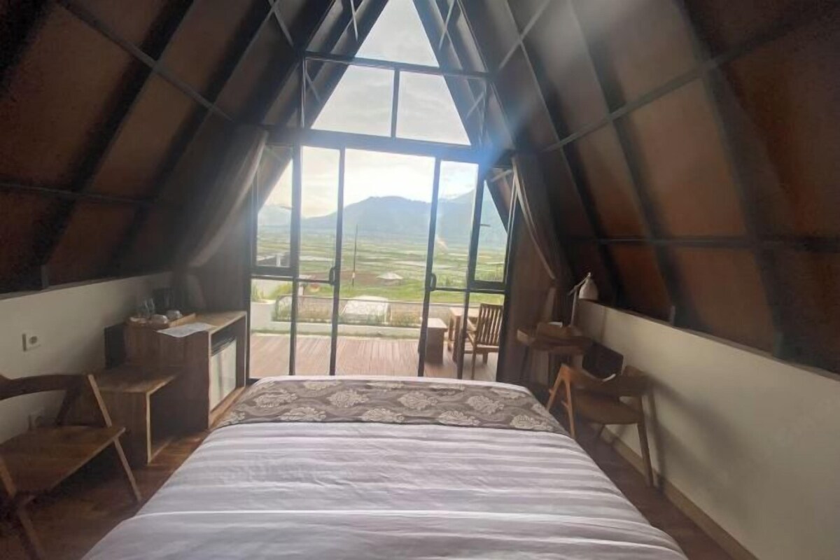 A bedroom with a large bed, a desk, and a chair, with a view of a landscape through a large window.