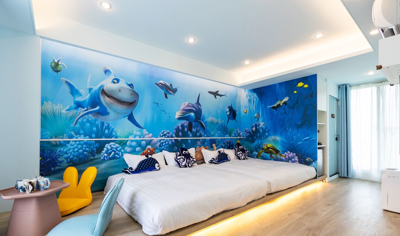 Un dormitorio con un gran mural de criaturas marinas en la pared, una cama con ropa blanca, una mesita con una tetera y una silla.