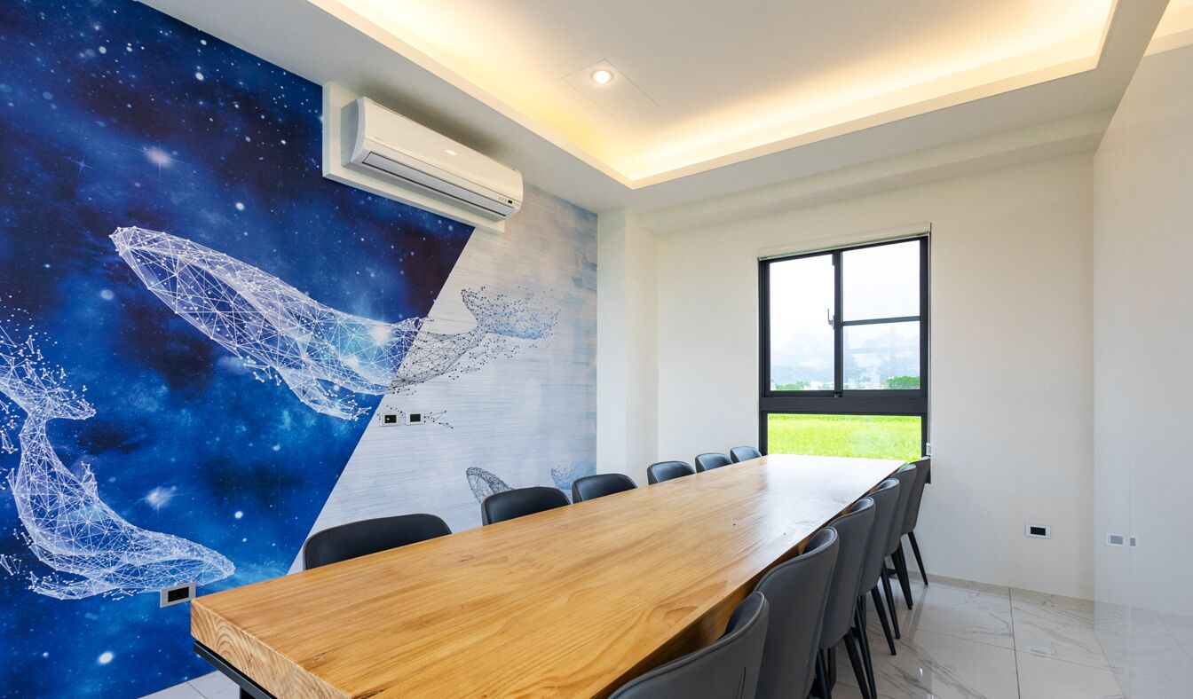 Un salón de conferencias con una mesa y sillas de madera, un amplio ventanal con vistas a zonas verdes y un mural en la pared con motivos espaciales.