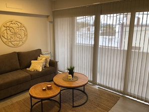 A living room with a brown sofa, two round wooden tables, a decorative wall piece, and a large window with vertical blinds. - Apartment first flor excellent location (Salta)