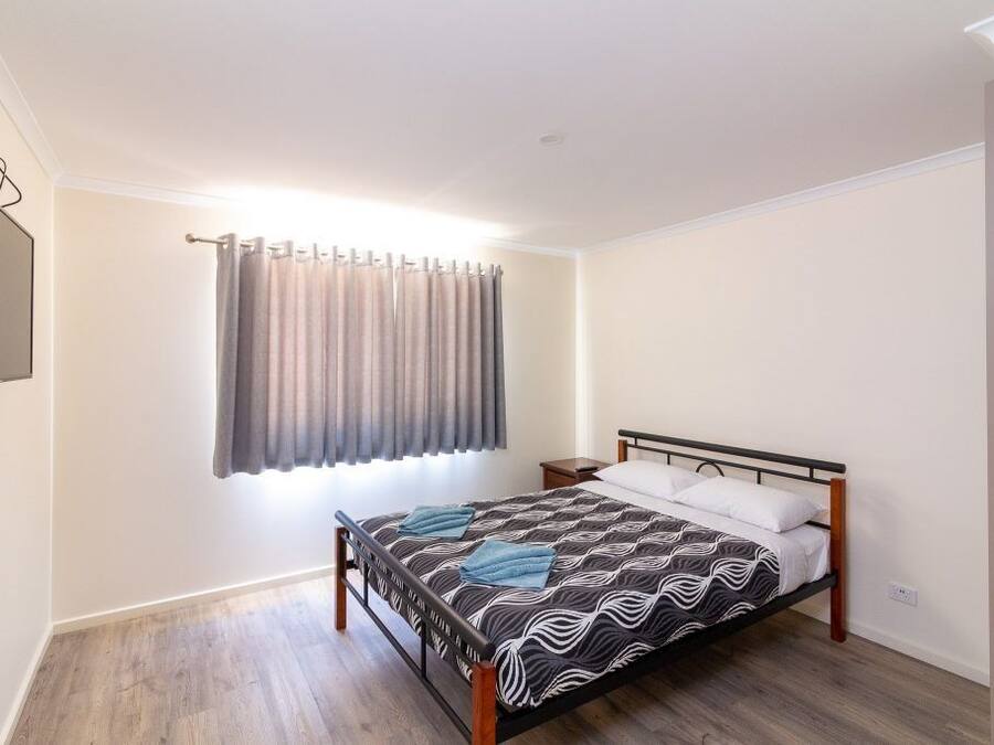 Een slaapkamer met een bed, een nachtkastje, een raam met gordijnen en een aan de muur bevestigde televisie.