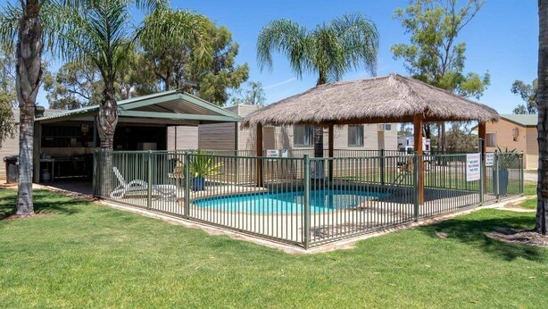 A pool area with a thatched-roof structure, surrounded by a metal fence, and a building with a green roof in the background. - Paringa Caravan Park (Paringa)