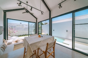 A modern room with a dining table, chairs, a sofa, and a large window overlooking a pool. - Villa Cha Cha  (Canggu)