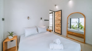 A modern bedroom with a large bed, a wooden side table with a plant, a mirror, and a view of greenery outside. - Villa Cha Cha  (Canggu)