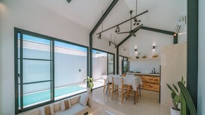 A modern interior with a dining area, kitchen, and a large window overlooking a pool. - Villa Cha Cha  (Canggu)