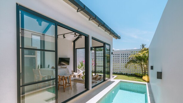 A modern house with a glass-enclosed patio, a swimming pool, and a view of a white fence and palm trees. - Villa Cha Cha  (Canggu)