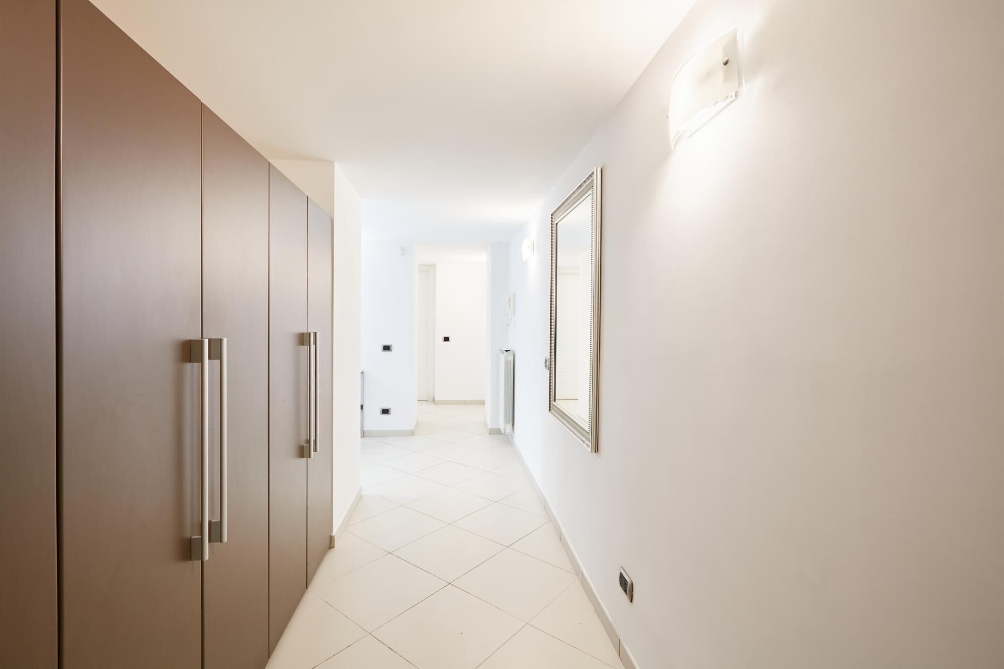 Ein Flur mit modernem Design, der einen langen Korridor, eine Tür am Ende und einen Bilderrahmenspiegel an der Wand zeigt.