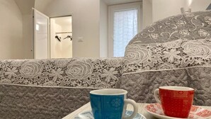 A room with a bed featuring a lace-patterned headboard, a wooden table with two cups, and a view of a hallway with a door and a window. - Grandma Lucia's house (Monopoli)