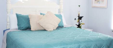 A neatly made bed with a teal bedspread and two pillows, a white headboard, and a framed picture on the wall.
