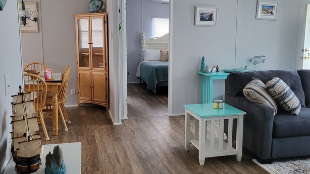 Un vaste espace de vie avec un canapé foncé, une table basse de couleur turquoise et un plancher en bois. On y trouve également un coin repas avec une table et des chaises en bois, ainsi qu’un meuble avec des portes en verre. On aperçoit un lit avec une tête de lit blanche à l’arrière-plan.