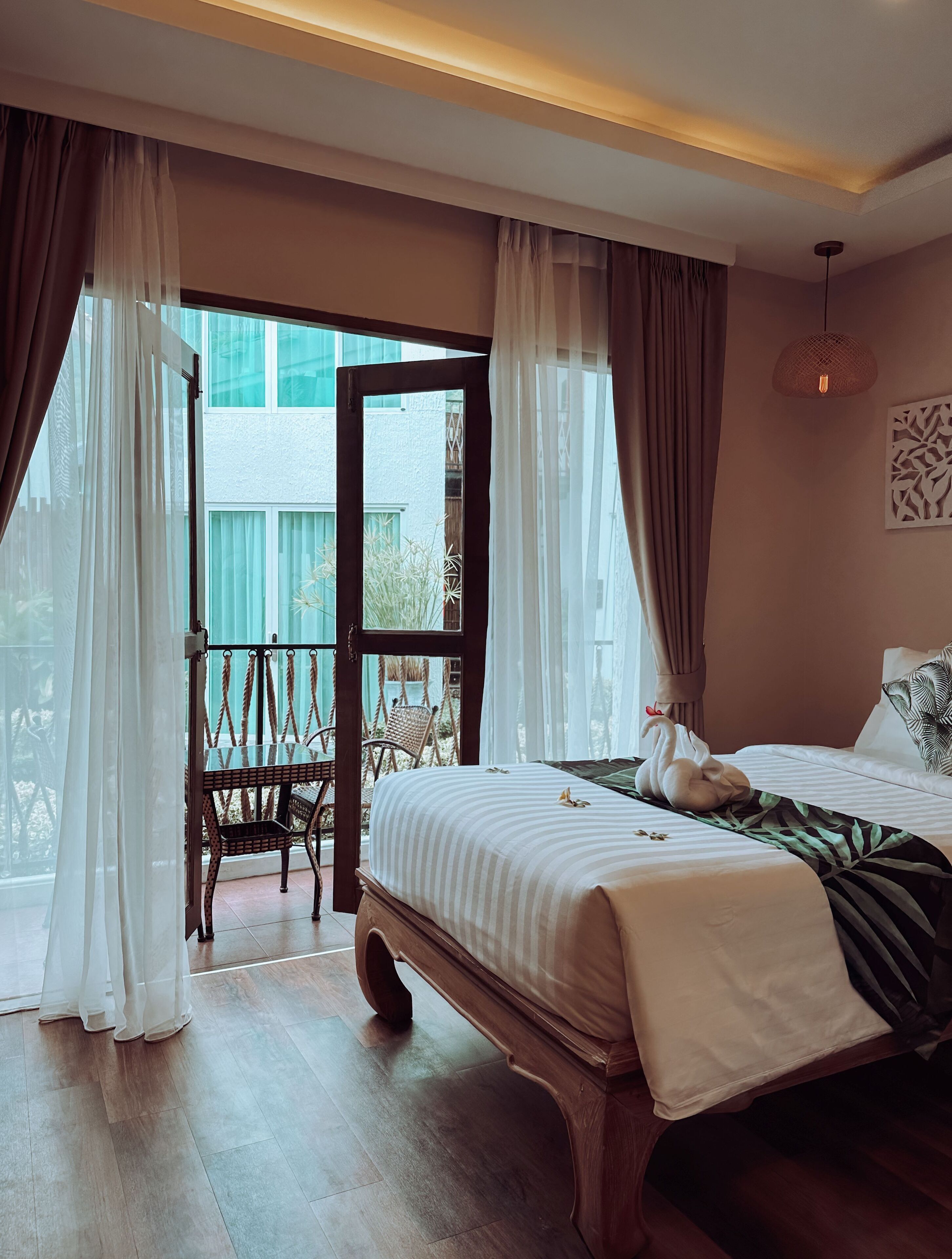 Habitación de hotel con una cama grande, un banco de madera, un balcón con sillas de mimbre y vistas a edificios.