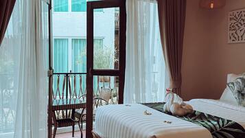 Habitación de hotel con una cama grande, un banco de madera, un balcón con sillas de mimbre y vistas a edificios.