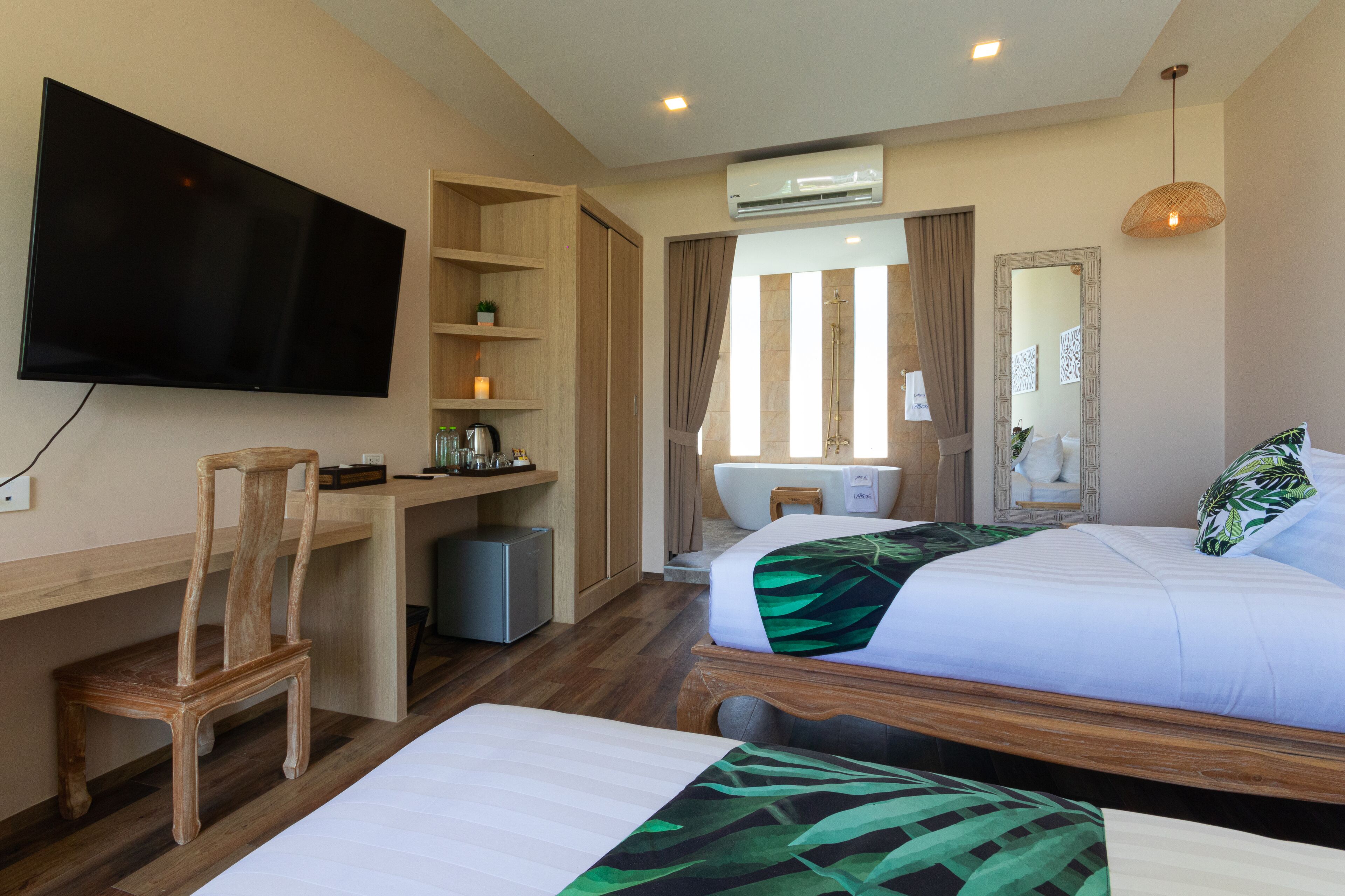 A hotel room with a flat-screen TV, a desk with a chair, two beds, a shower area, and a large mirror.