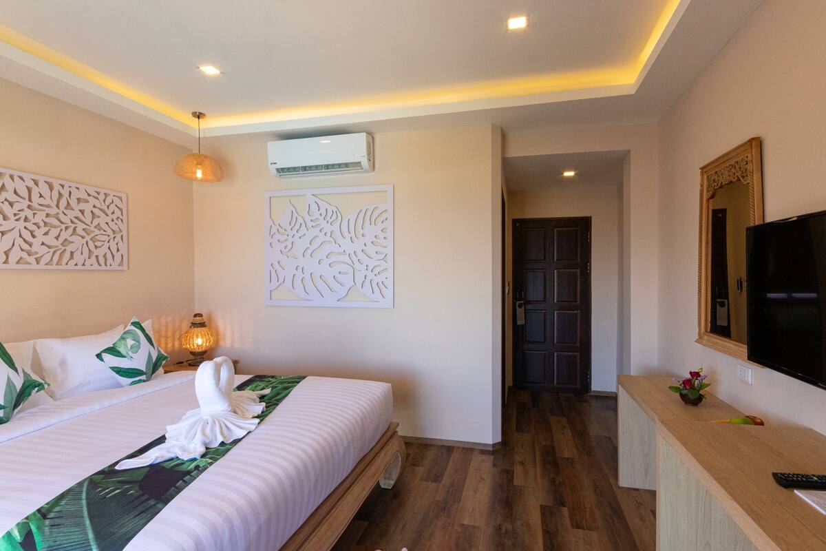 A hotel room with a bed, a bedside lamp, a wall-mounted air conditioner, a decorative wall panel, a wooden floor, a TV, and a small plant on the dresser.