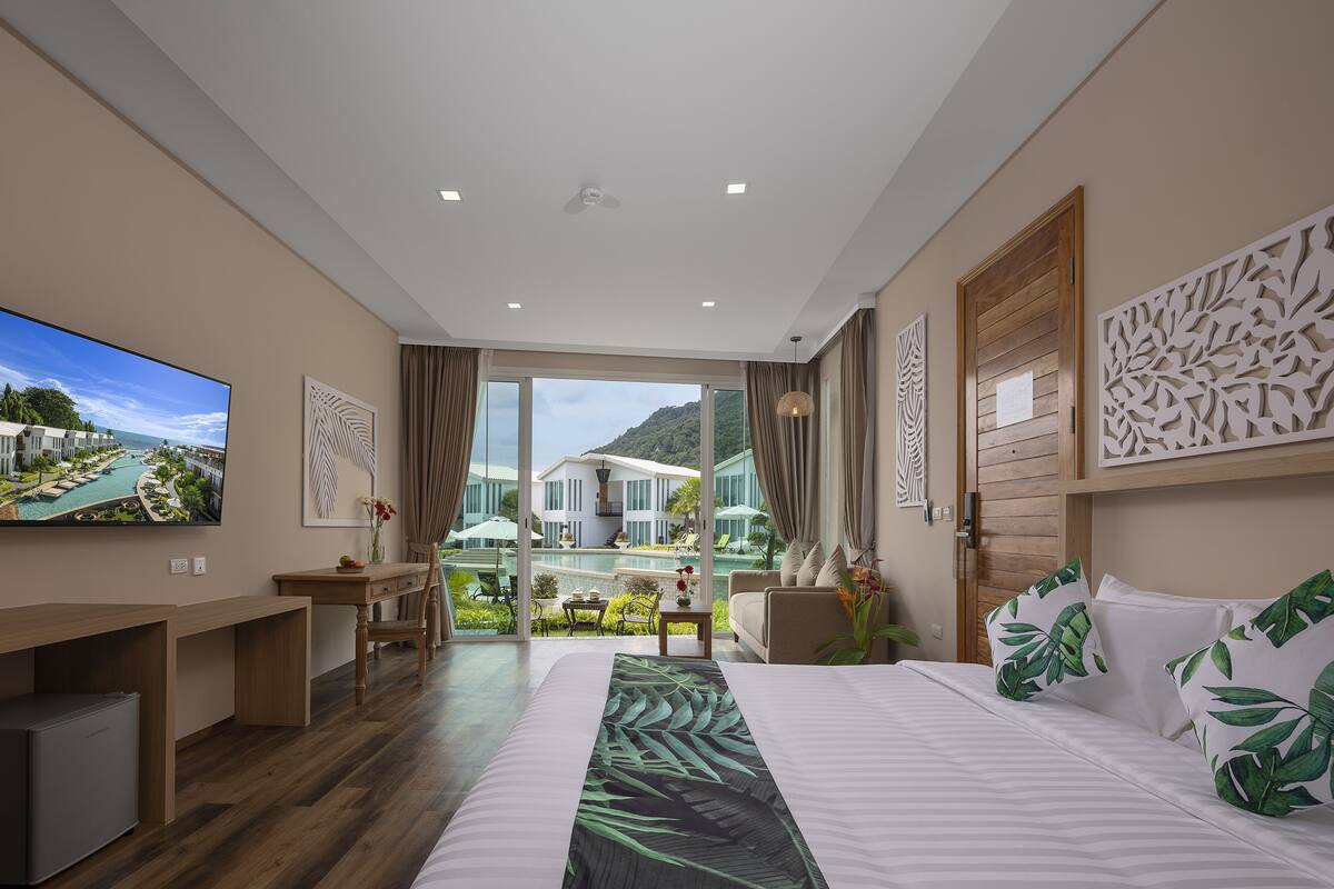 A modern hotel room with a large bed, a desk, a television, and a view of a pool and mountains.