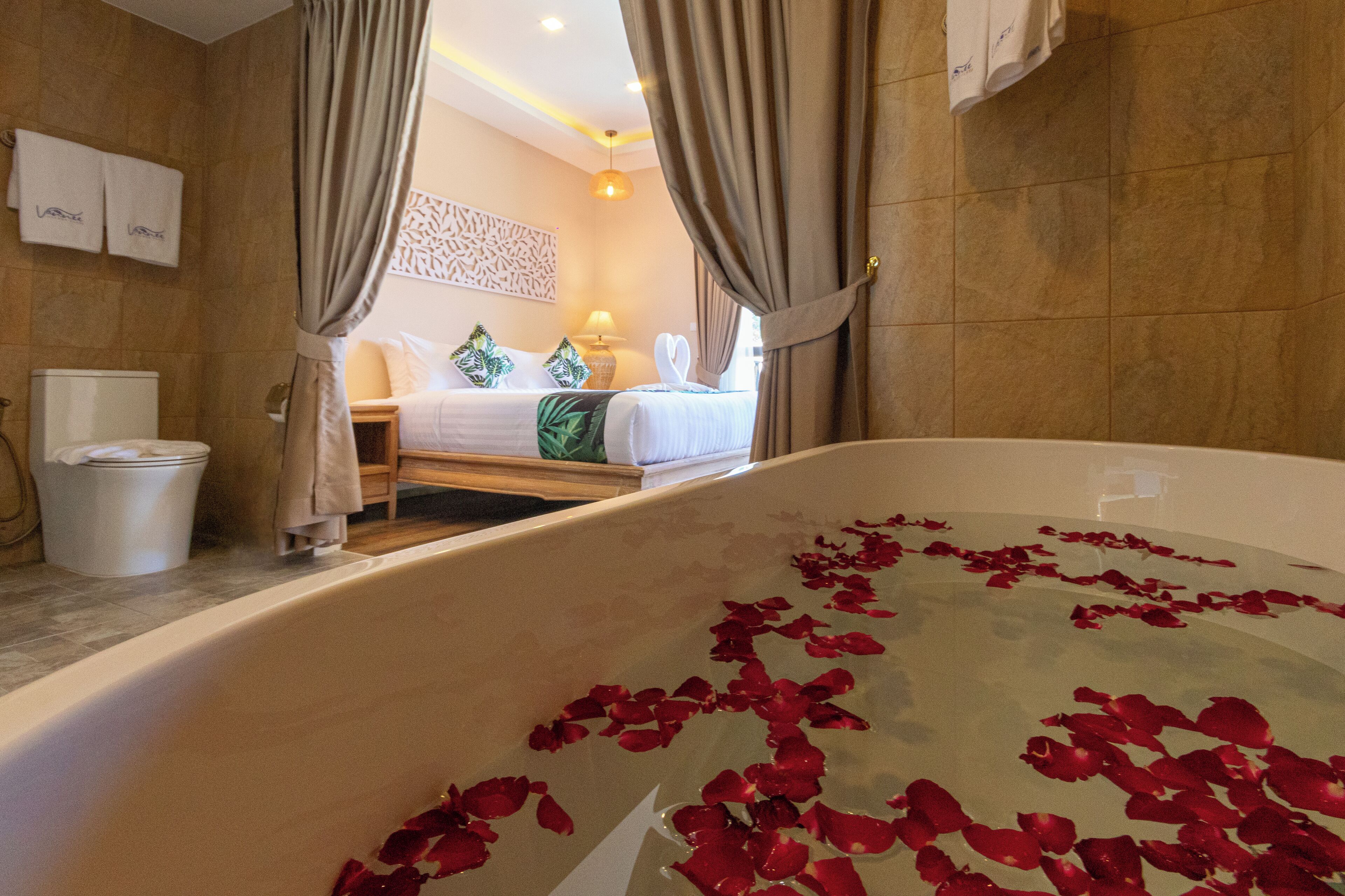 A bathroom with a bathtub filled with rose petals, a toilet, and a bed with a nightstand.