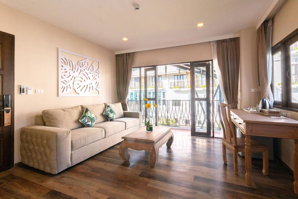A living room with a sofa, coffee table, and dining table. There is a large window with curtains, a decorative wall piece, and a view of a balcony.