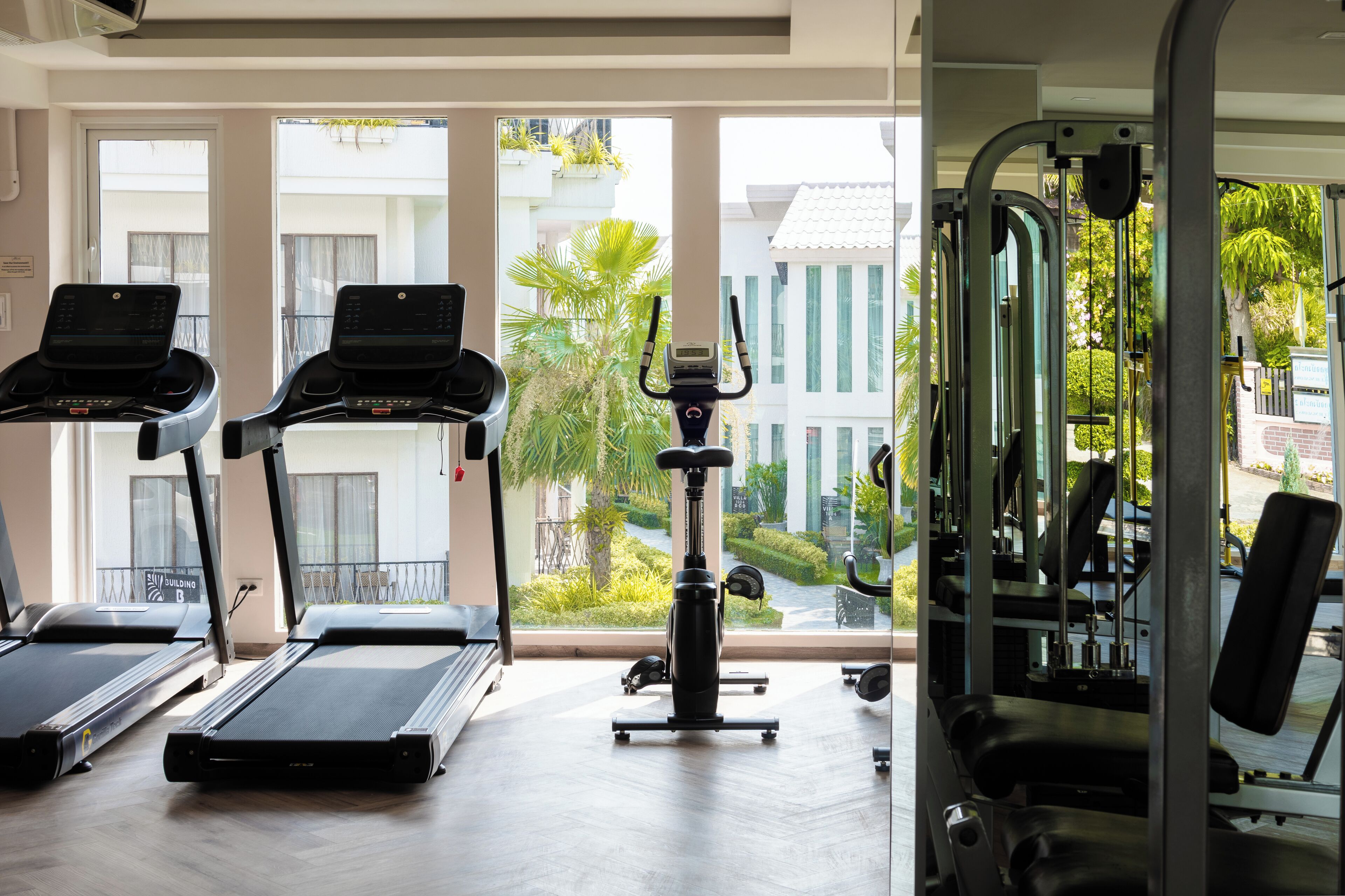 Un gimnasio con cintas de correr, una bicicleta estática y máquinas de pesas. Cuenta con ventanas amplias que ofrecen vistas a un edificio y zonas verdes.